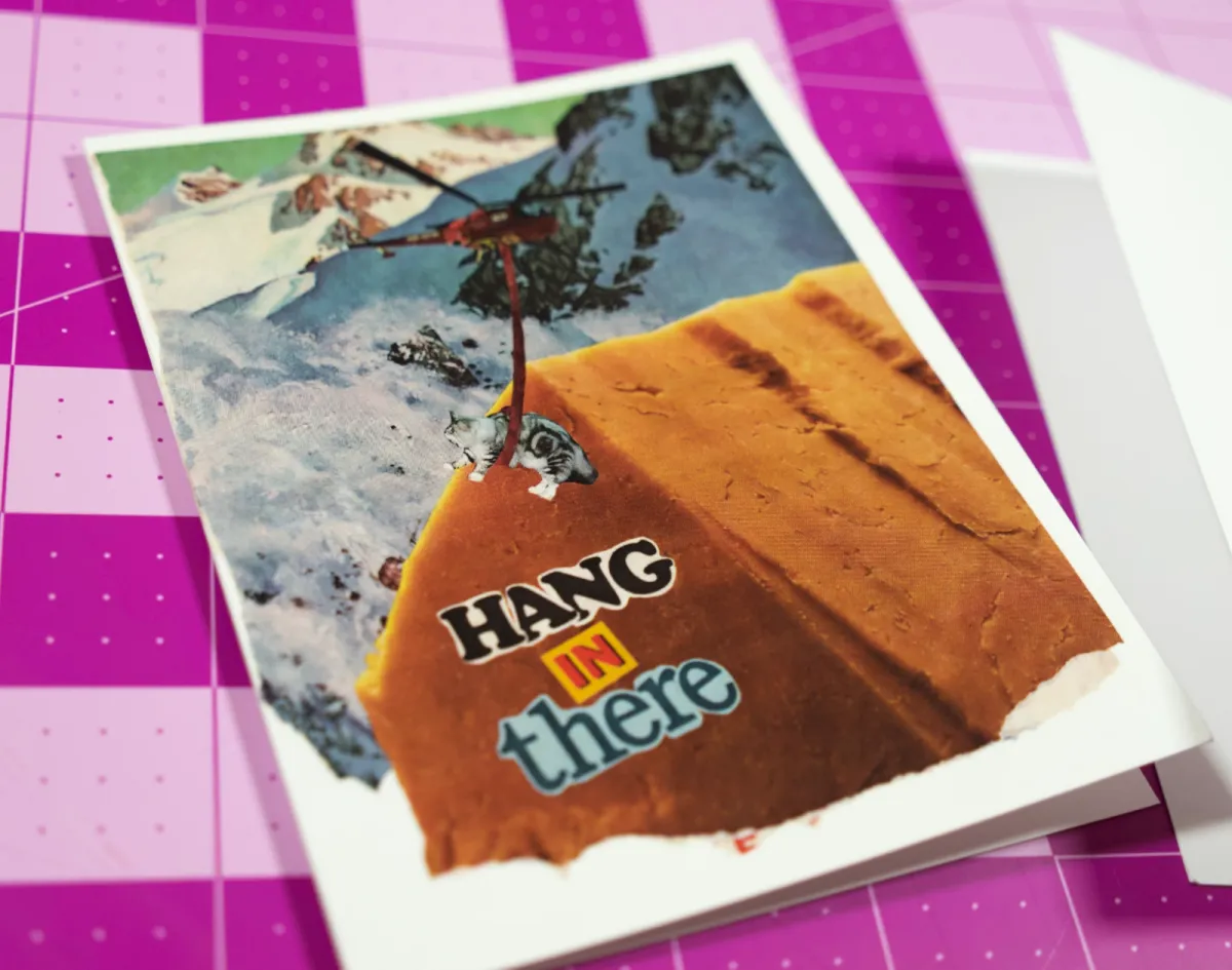 A close-up of the printed Hang in There greeting card with its white envelope on a pink cutting mat.